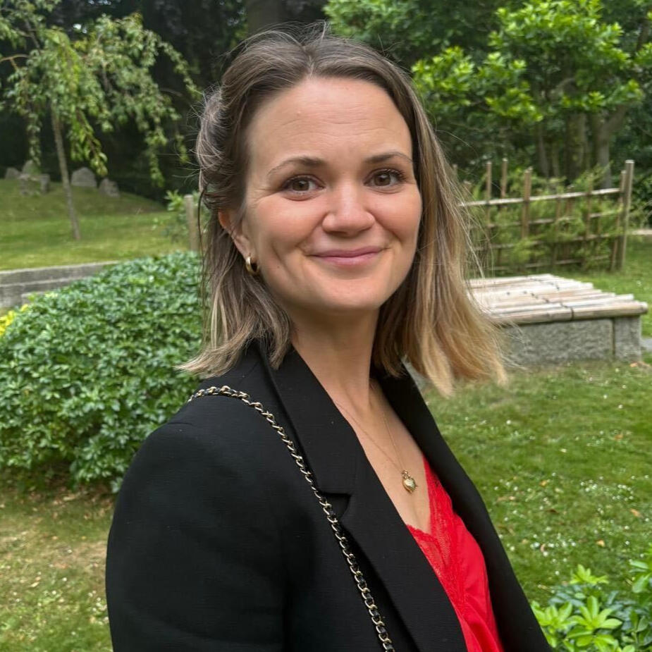 Alex, a white woman with light brown hair in a sharp bob, is standing in a garden wearing a red top and black blazer.
