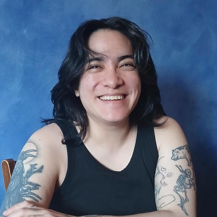 Rho, a mixed East Asian person, smiles in front of a blue background. They are wearing a black tank top that shoes their tattooed arms