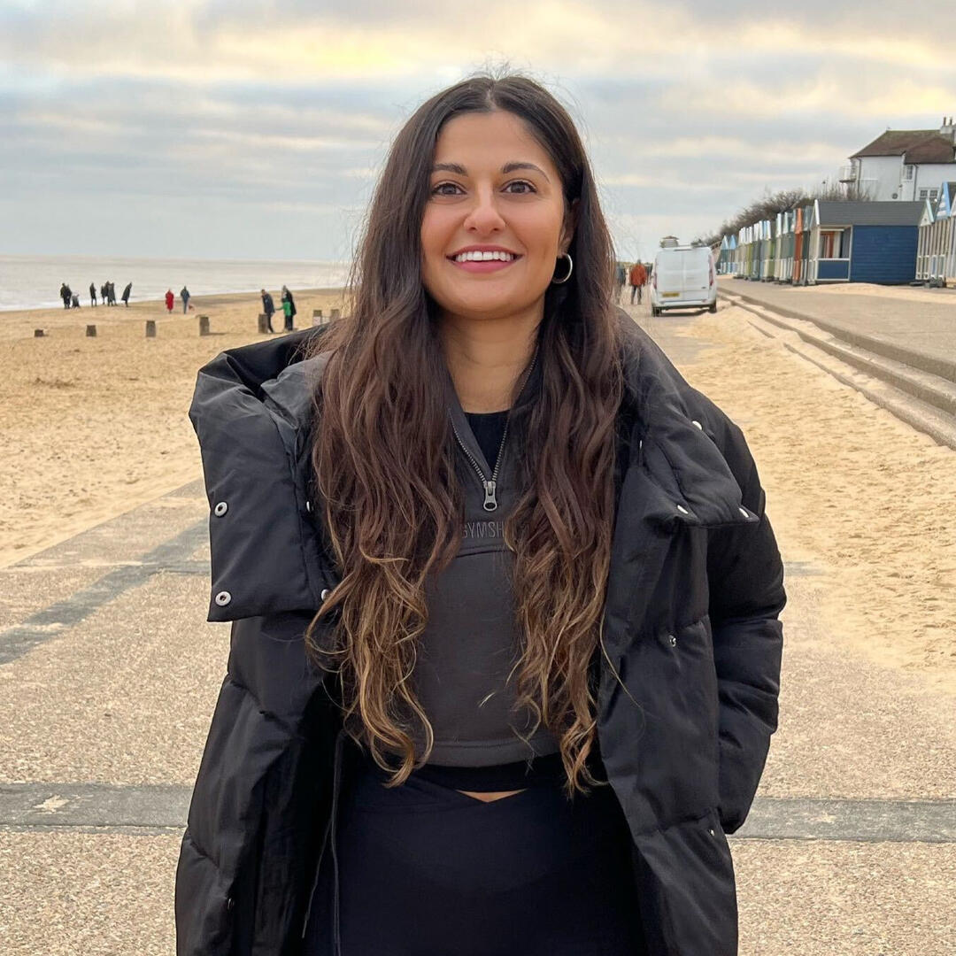 Aysha, a brown-skinned woman with long wavy black hair, smiles. She has her hands in her pockets of a warm black puffer jacket and is standing on a beach on an overcast day