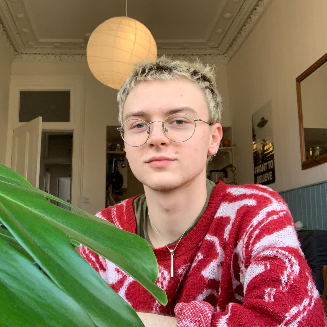 Levi sitting next to his monstera plant and wearing a red jumper. They are white and have short blond hair and glasses.