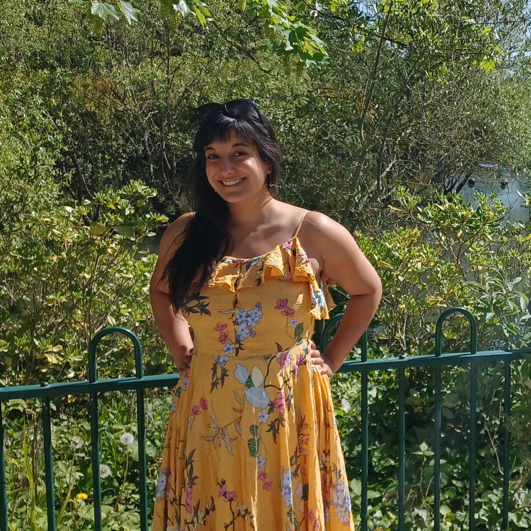 Michelle, a brown-skinned woman with very long black hair, is standing with her hands on her hips in front of a backdrop of trees. She is wearing a yellow sundress, has a pair of sunglasses on her head and is smiling.