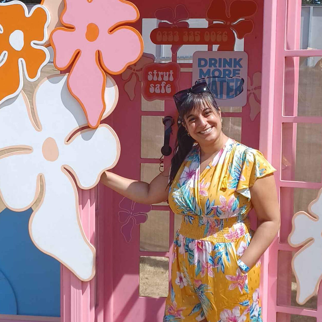 Michelle, a brown-skinned woman with very long black hair tied back in a ponytail, is posing in a bright pink Strut Safe branded phone box. Michelle is wearing a floral yellow jumpsuit.