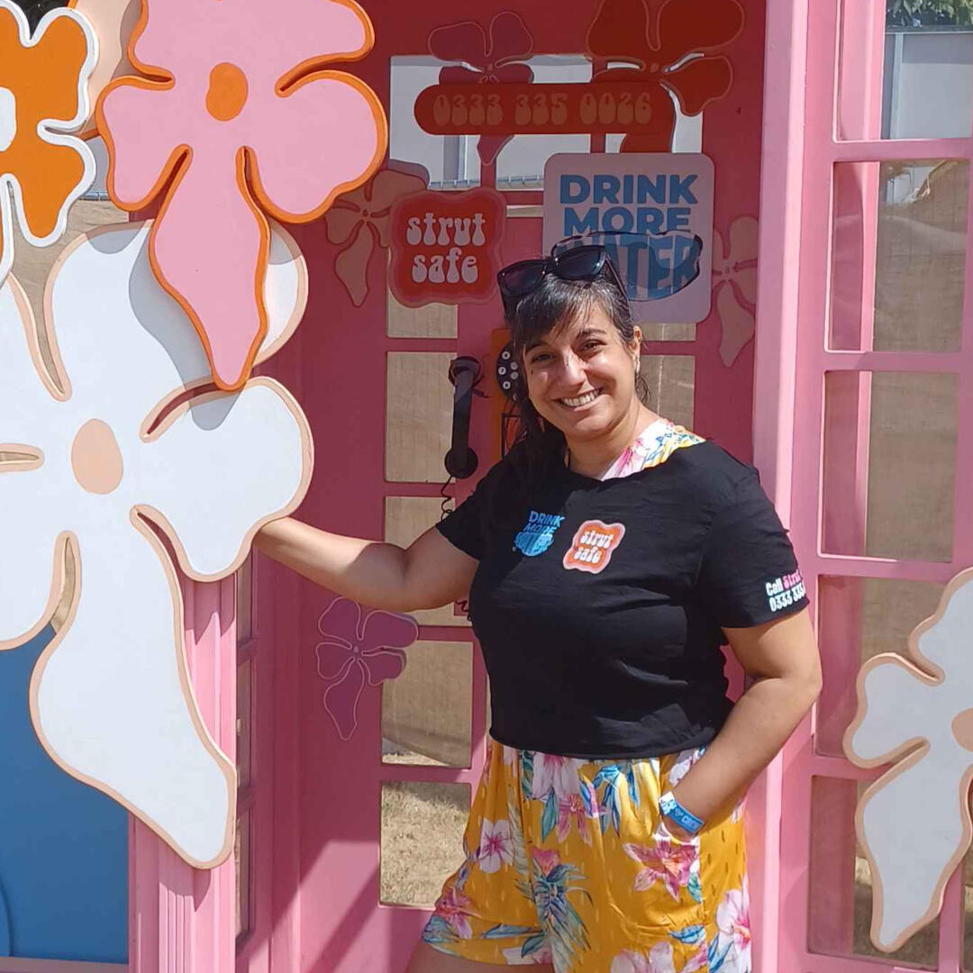 Michelle, a brown-skinned woman with very long black hair, is posing in a bright pink Strut Safe branded phone box. Michelle is wearingyellow floral shorts and a black t-shirt with the Strut Safe logo on.