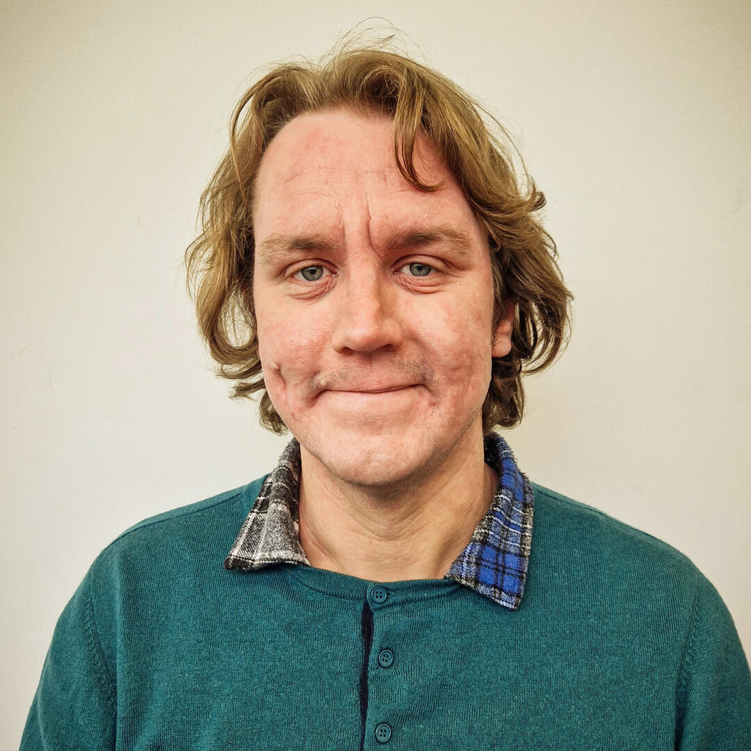 Diar, a white person with long blond hair, is sitting outdoors next to a carved wooden goose. They wear a patterned sunhat and a black and white baseball shirt.