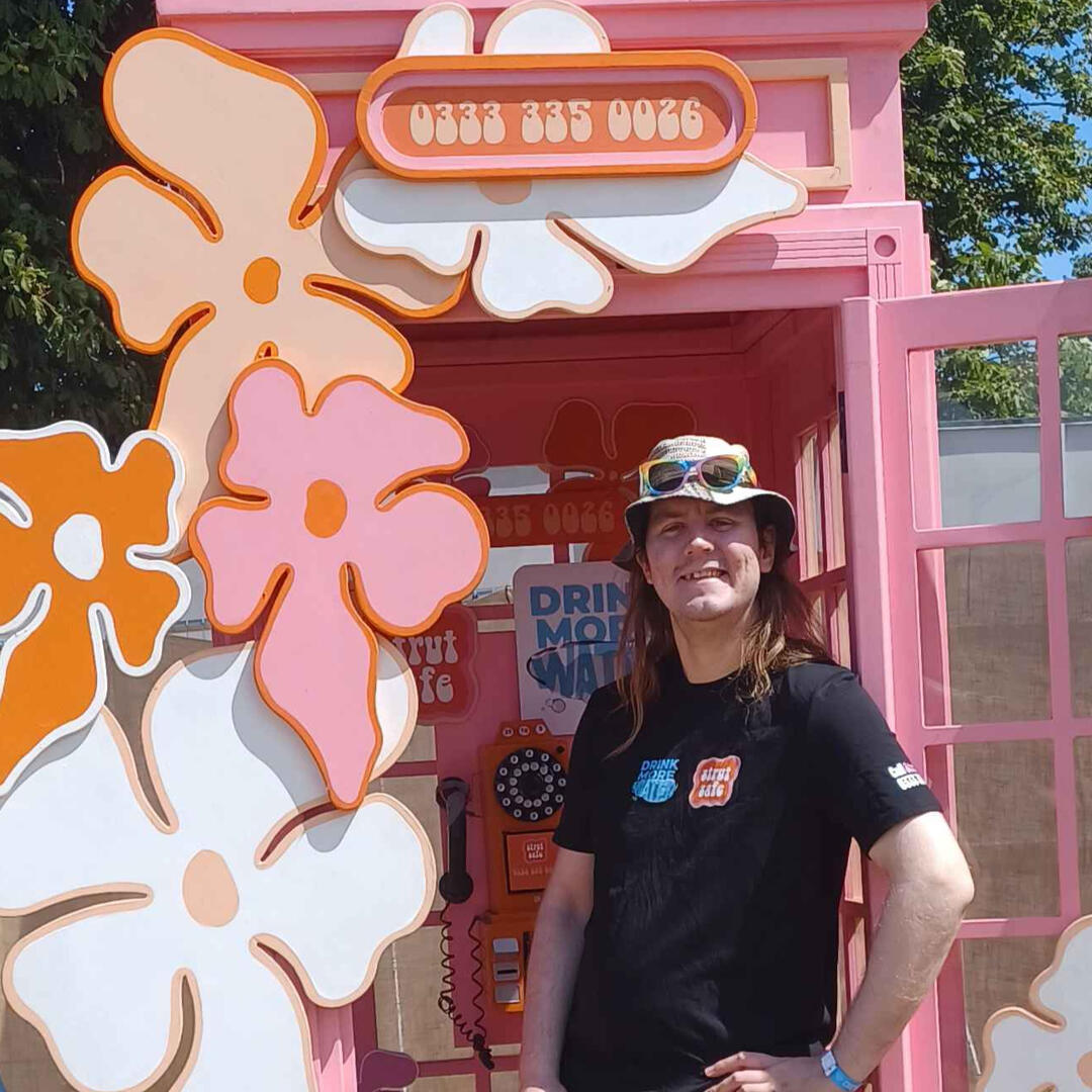 Diar, a white person with long blond hair, is standing in front of a bright pink Strut Safe branded phone box. They are wearing a sunhat and a black t-shirt with the Strut Safe logo on it.