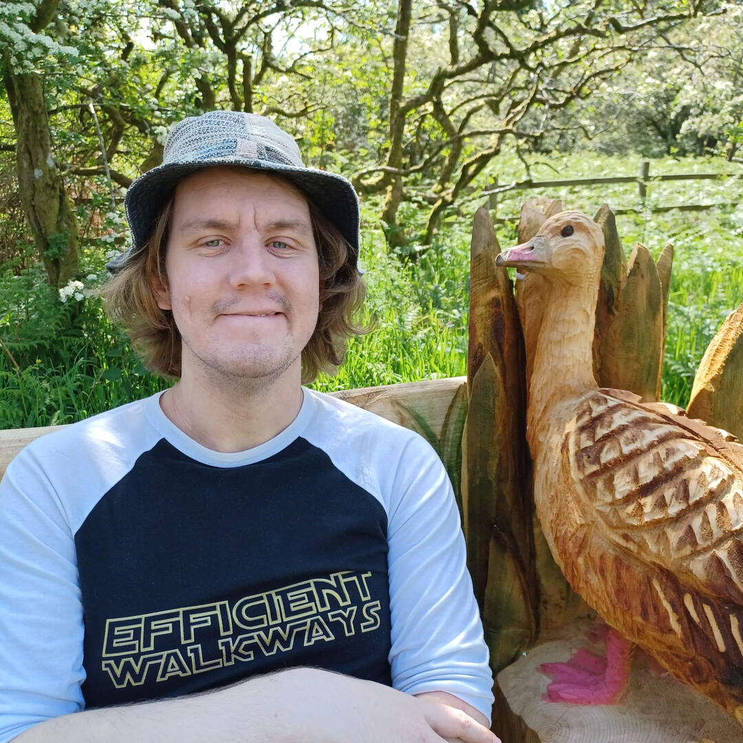 Diar, a white person with long blond hair, is sitting outdoors next to a carved wooden goose. They wear a patterned sunhat and a black and white baseball shirt.