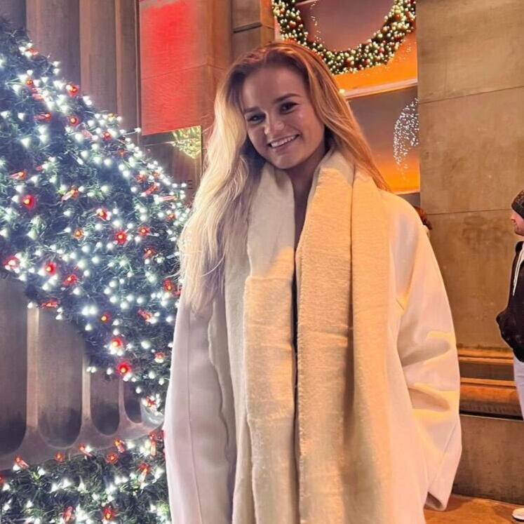 Kimberley, a white woman with long blonde hair, is smiling and standing in front of a large wreath decorated with fairy lights. She wears a warm cream-coloured coat.
