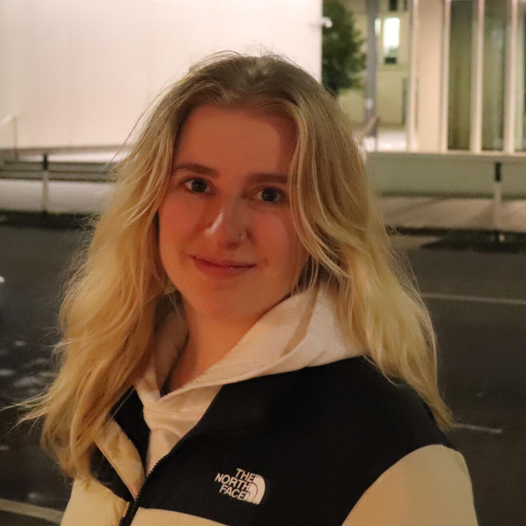 Katie, a white woman with long blonde hair, smiling and standing on a street at night wearing a black and white coat.